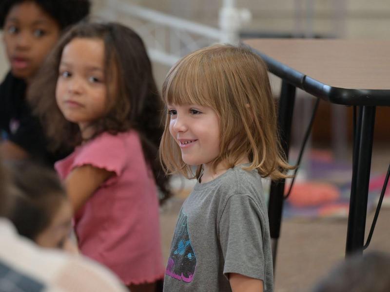 Young girl smiling in a classroom with other children in the background.