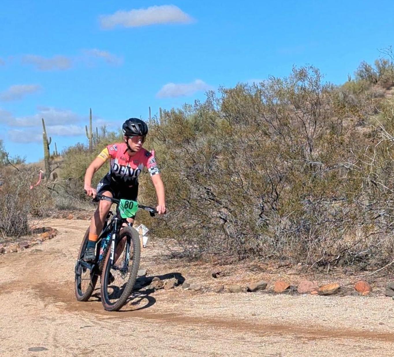A cyclist in action on a dirt path surrounded by desert vegetation under a blue sky.