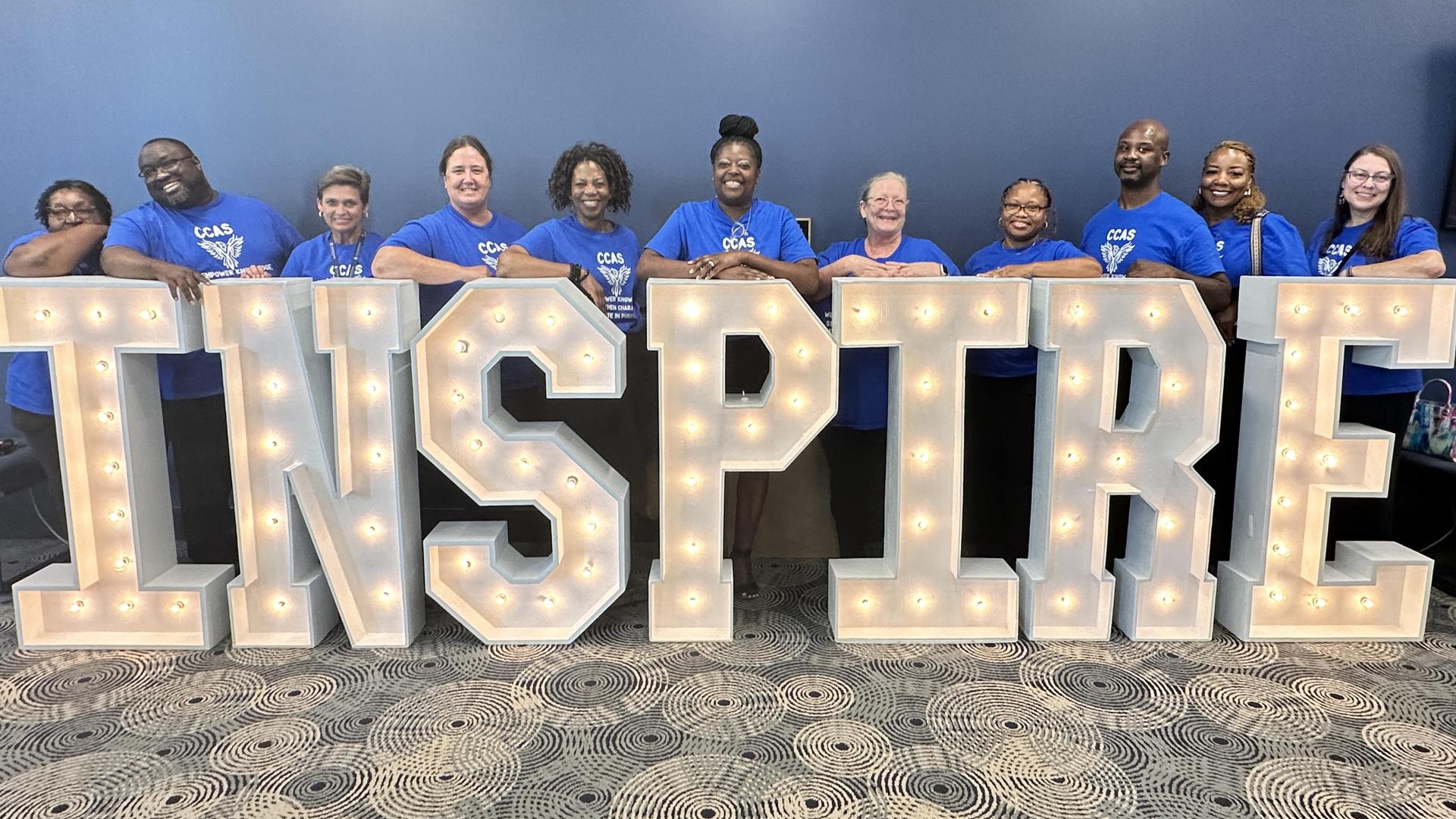 Group of people in blue shirts standing behind a large, illuminated sign that says 'INSPIRE'.