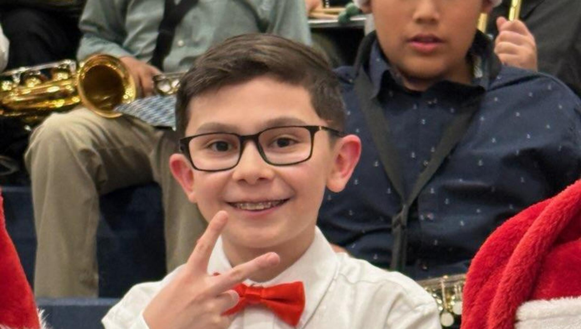 Smiling boy in a white shirt and red bow tie making a peace sign, surrounded by Christmas hats.