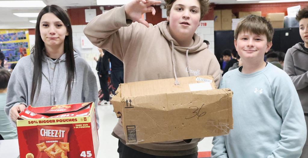 A female student standing holding a Cheez-It box.   A male student standing in the middle holding a cardboard box.  A boy standing holding a pillow case with books in it.
