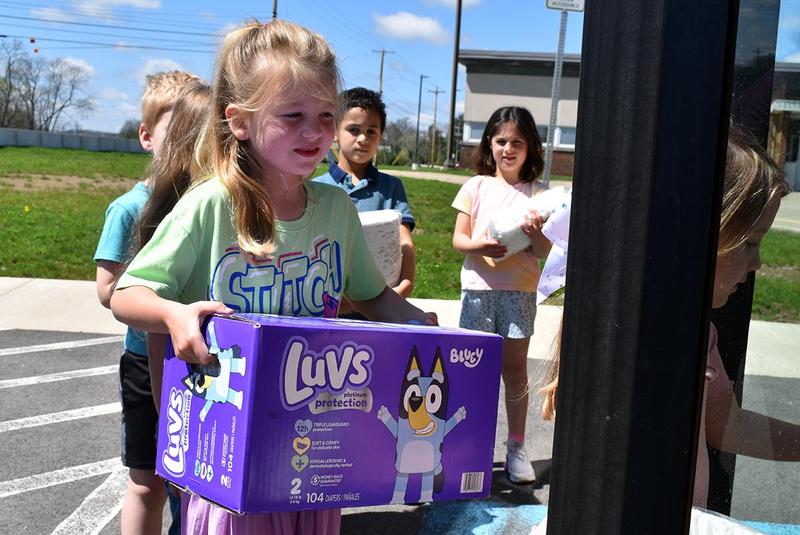 Children carrying boxes of diapers outside a building on a sunny day.