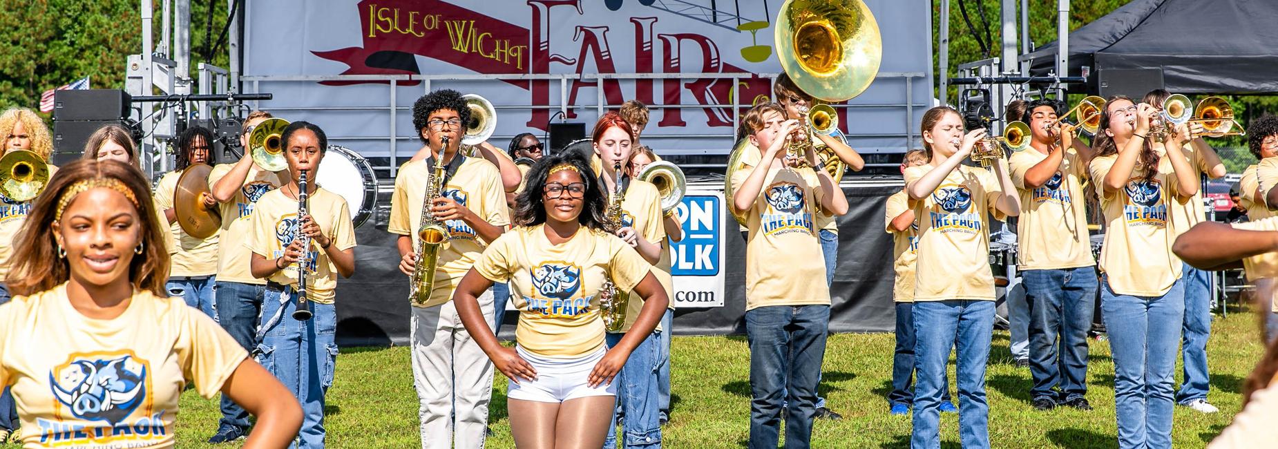 Marching band members playing instruments at an outdoor fair, with dancers in front.