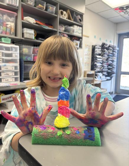 A girl displays her colorful art project and painted hands, smiling at the camera in a classroom.