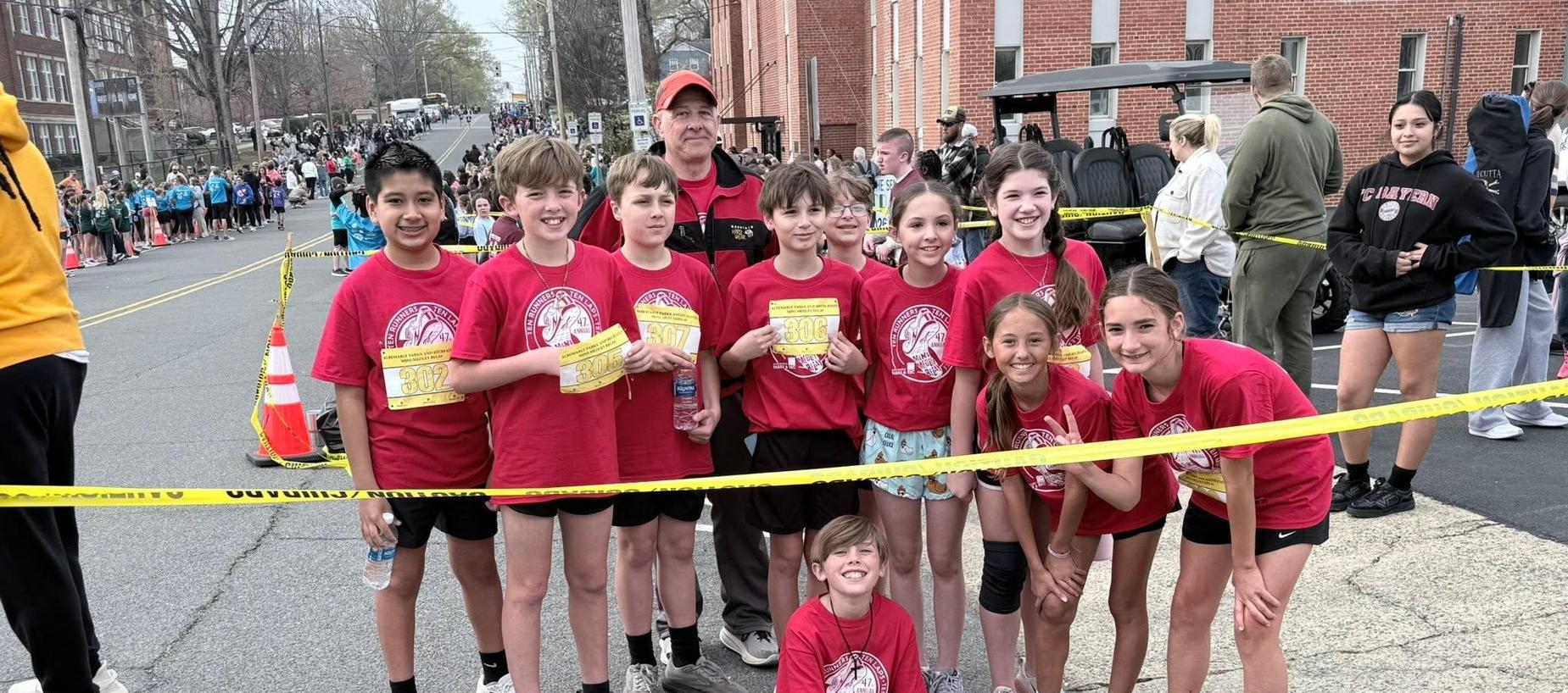 Group of children in red shirts posing at a running event with spectators in the background.