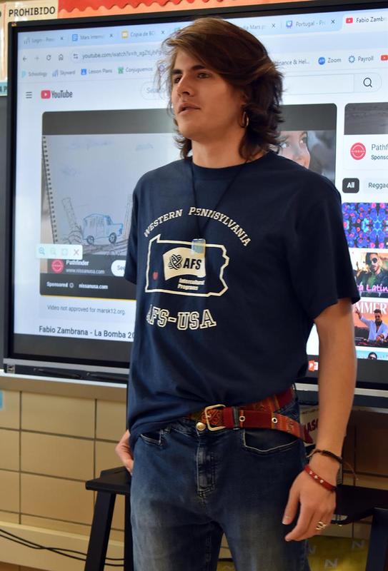 A student speaks in front of a screen showing a YouTube video, wearing a dark t-shirt.