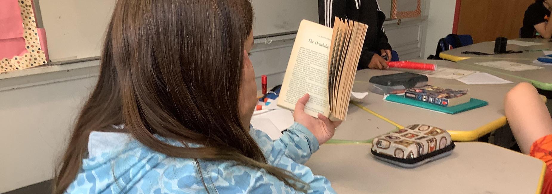 Student in a blue jacket holding a book at a classroom table.