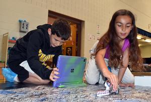 Two kids use a tablet and a toy car on an educational mat.