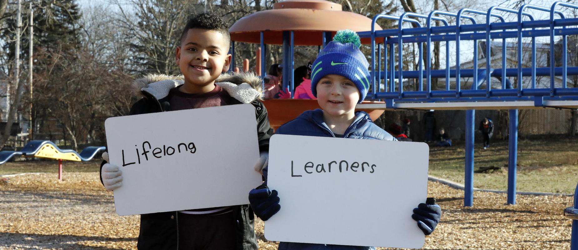 boys pose with lifelong learners sign on playground in their winter gloves and hats and coats