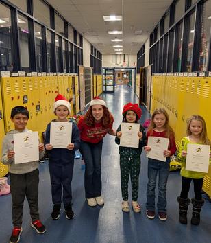 Five children and one adult in holiday attire holding certificates in a school hallway.