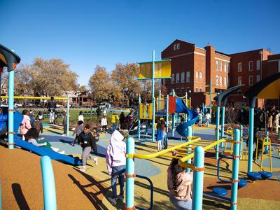 Children play on colorful playground equipment while others gather near a school building.