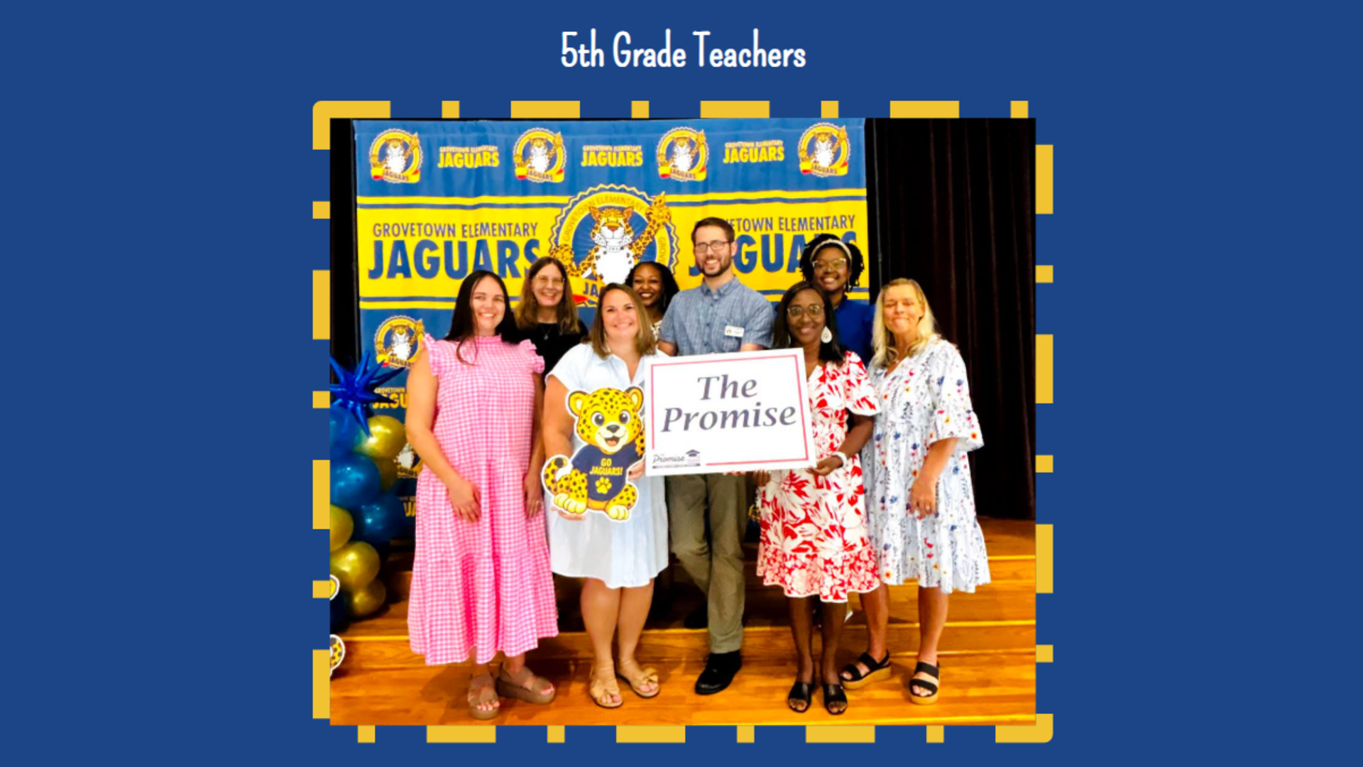 A group of eight teachers stand with a sign and a mascot, posing in front of a school banner.