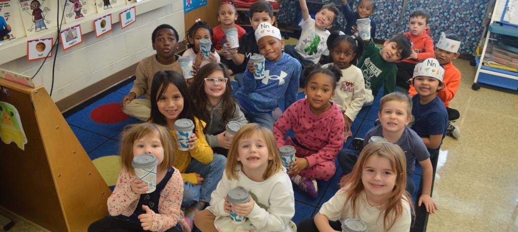 class of students sit on a brightly colored rug and raise their cups of hot chocolate
