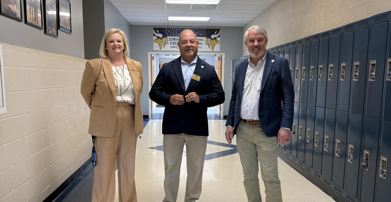 Three adults stand in a school hallway next to lockers, smiling at the camera.