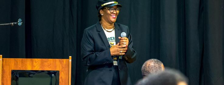 A speaker in a cap and blazer smiles while holding a microphone at an event.
