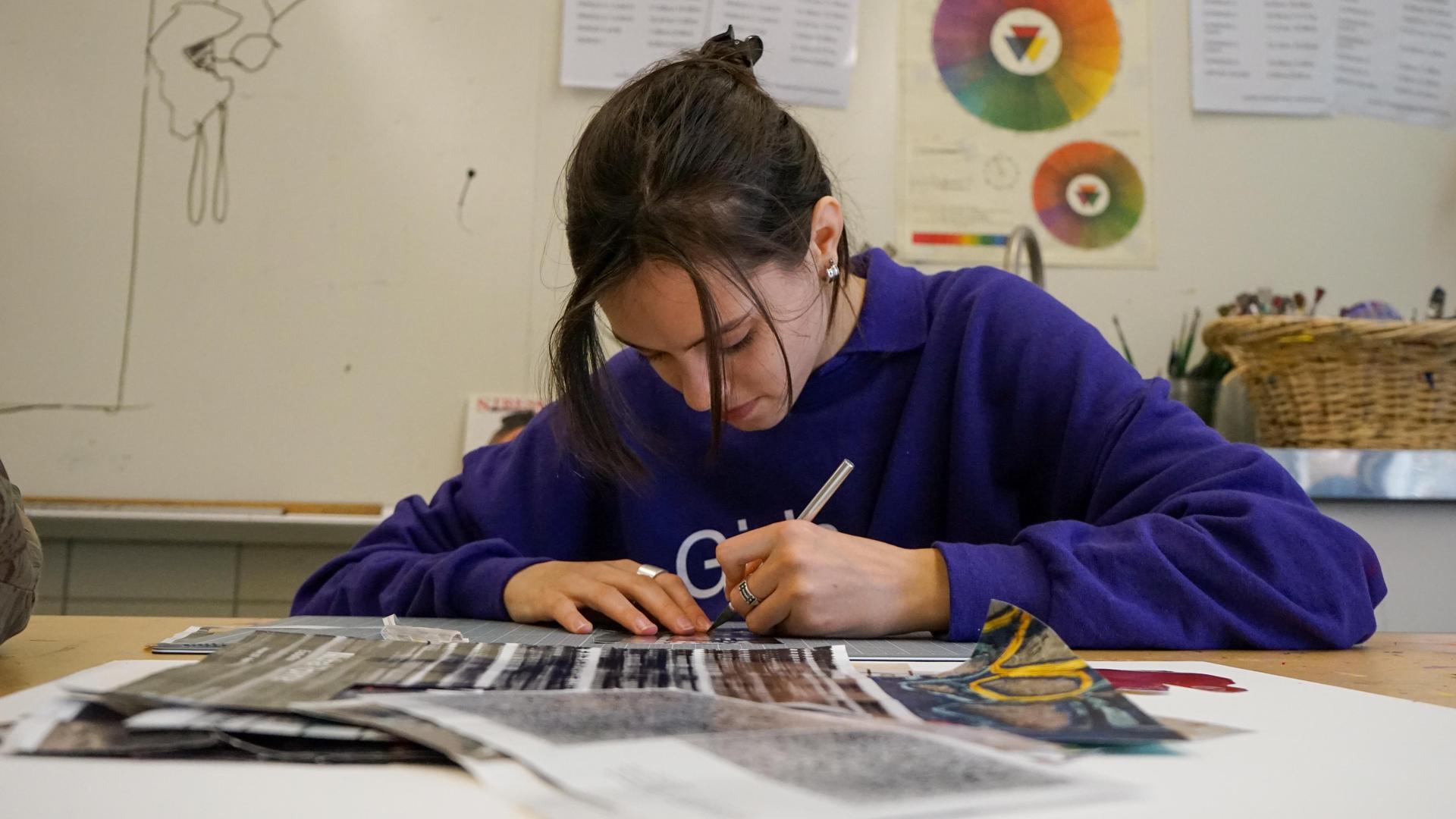 A student in a purple sweater sketches on a table filled with art supplies.