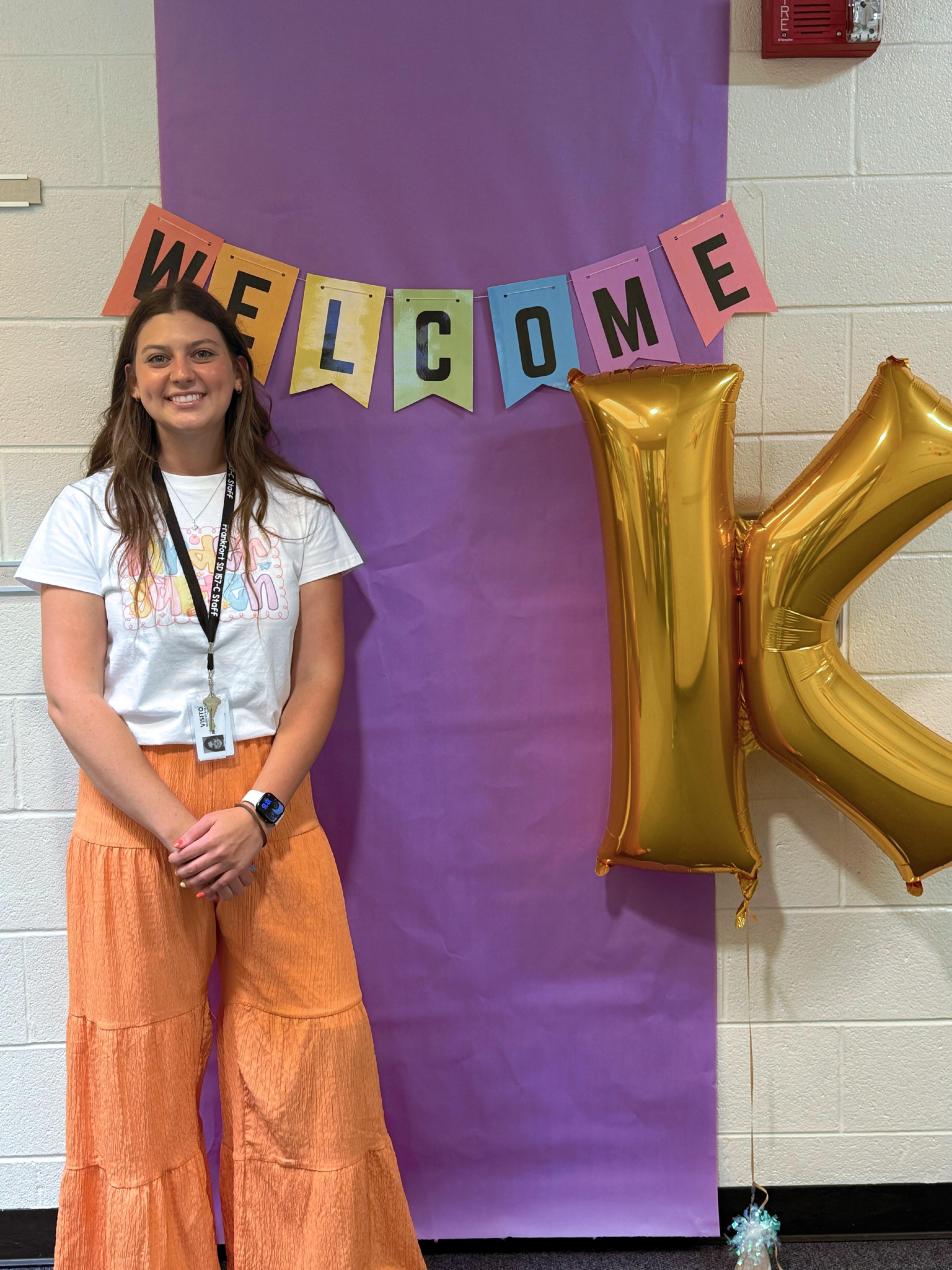 A smiling woman in a colorful shirt stands beside a 'Welcome' banner and a gold balloon.