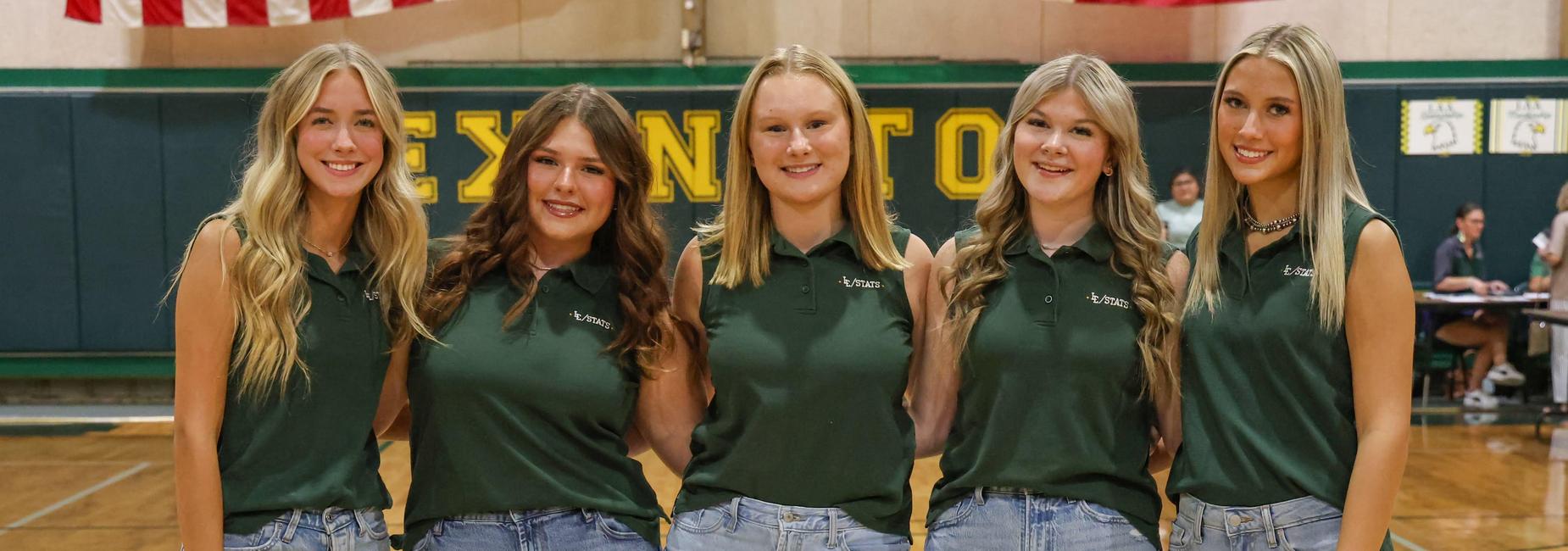 Five individuals in green tops and jeans posing together on a basketball court.