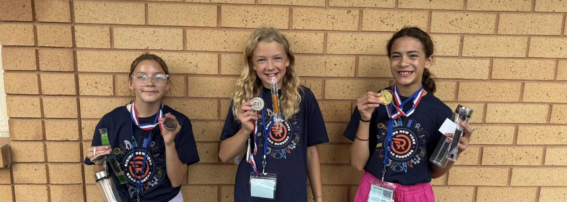 Three girls proudly displaying their medals and trophies against a brick wall.