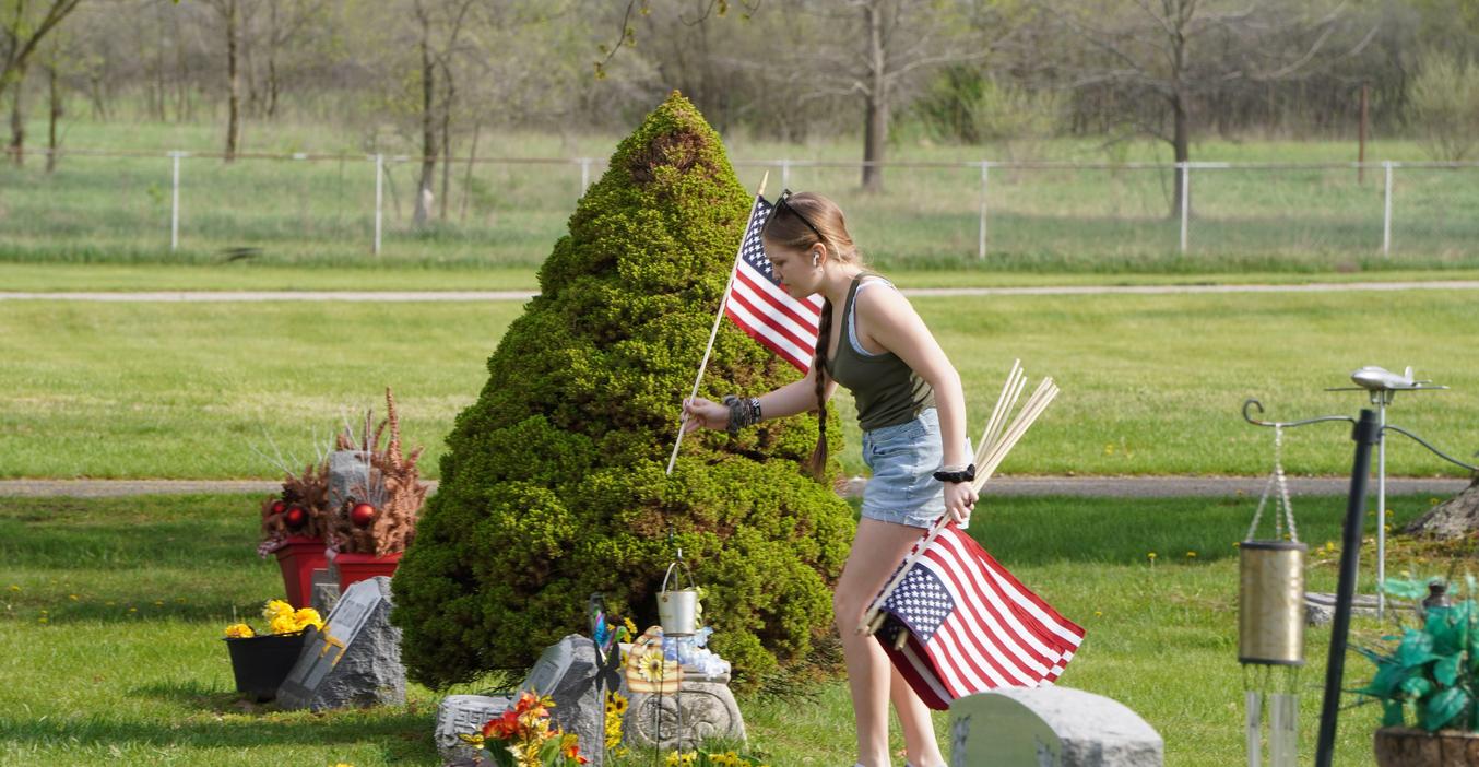 A young person placing flags at a grave site surrounded by flowers and a shrub.