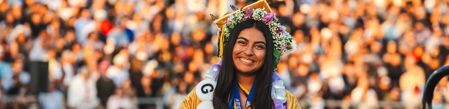 high school graduate in yellow cap and gown stands in front of a large crowd