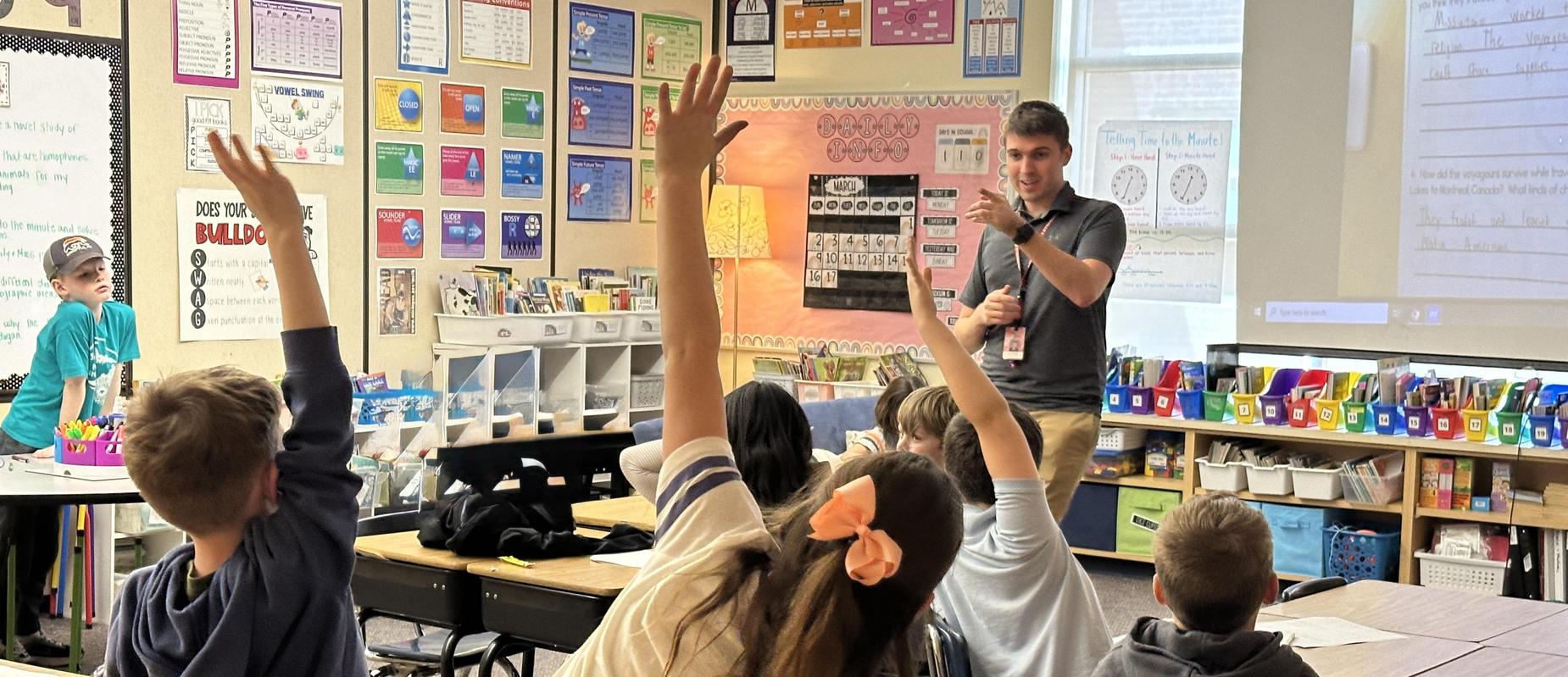 teacher in grey shirt smiles and points to child raising their hand in an elementary classroom with three hands in the air