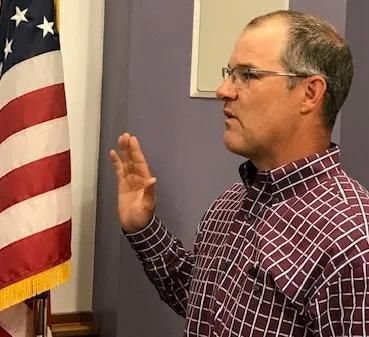 Man in a checkered shirt raises his hand near an American flag.
