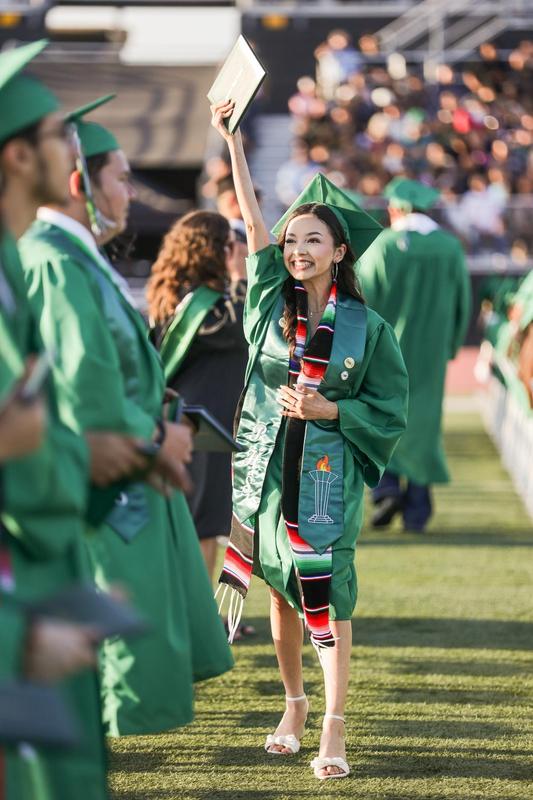A female Pioneer High School graduate lifts her diploma in the air, smiling to her family in the stands.