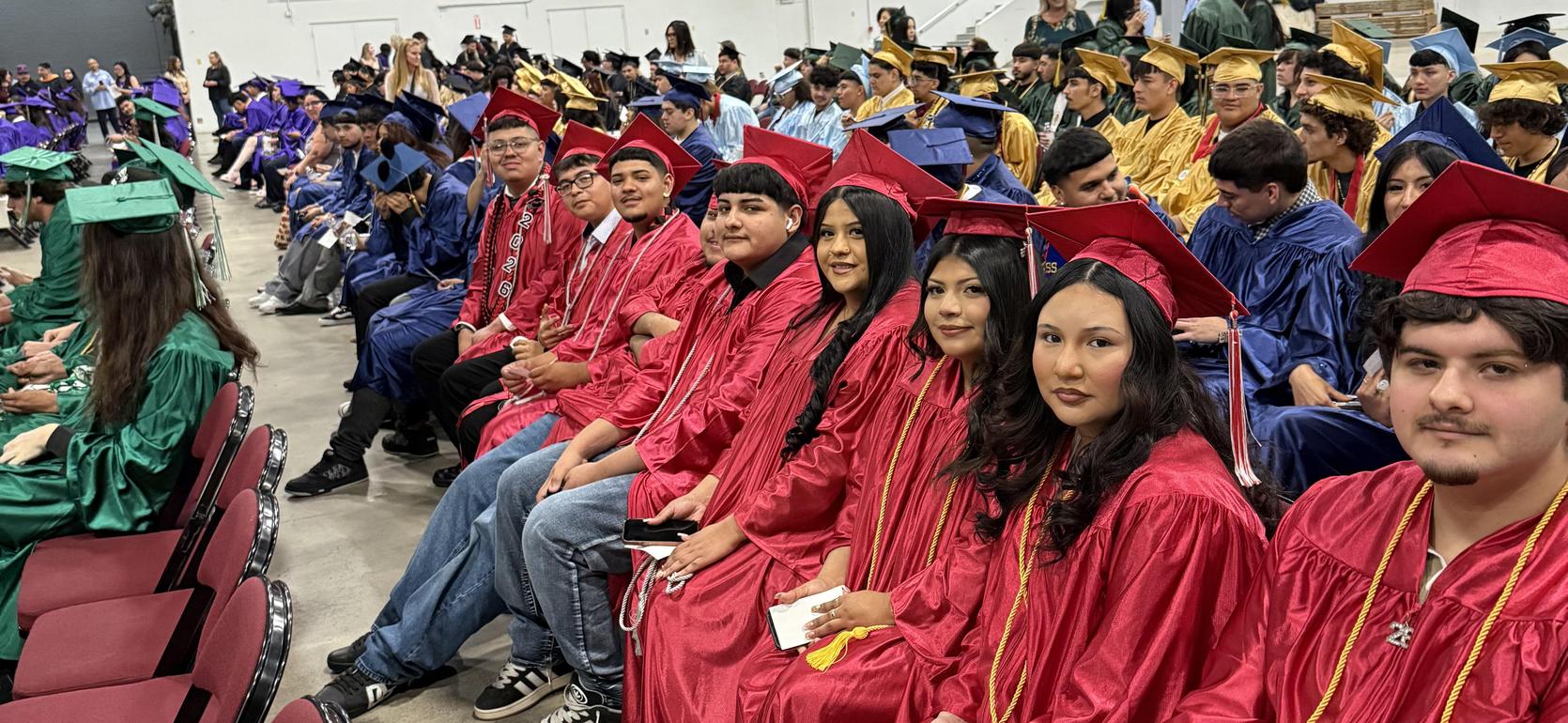 Graduates in red, green, and blue gowns seated in a hall, awaiting the ceremony.