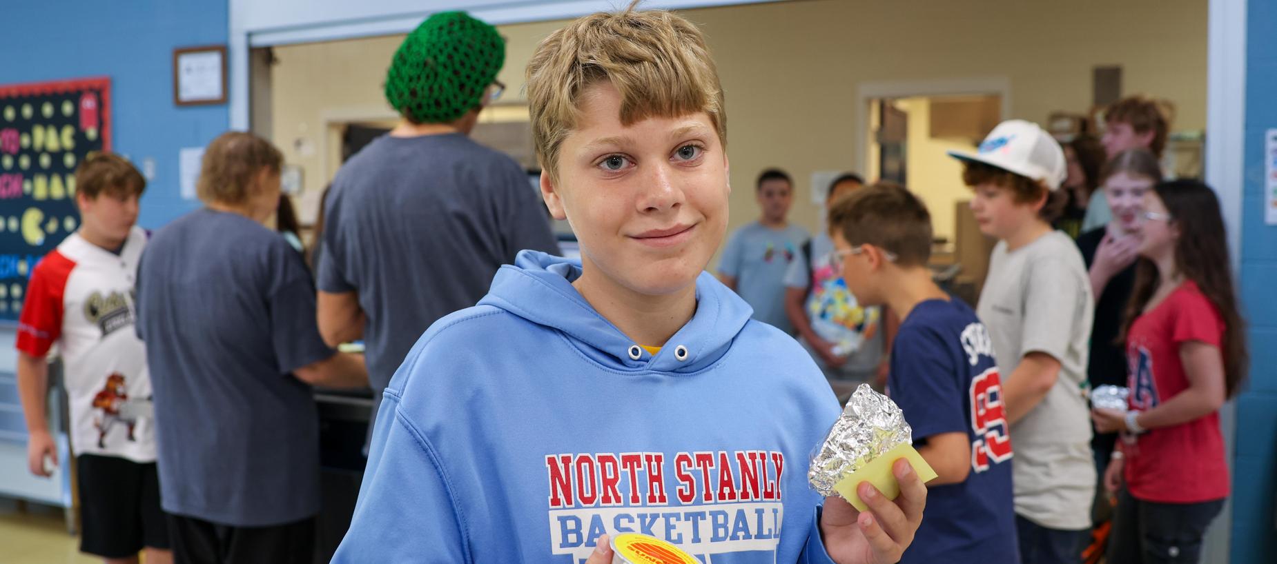 A boy in a blue hoodie holds a snack while smiling in a crowded cafeteria.