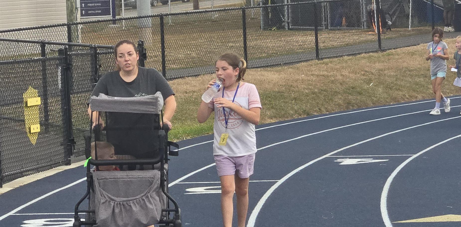 A woman pushing a stroller alongside a girl drinking water on a track.