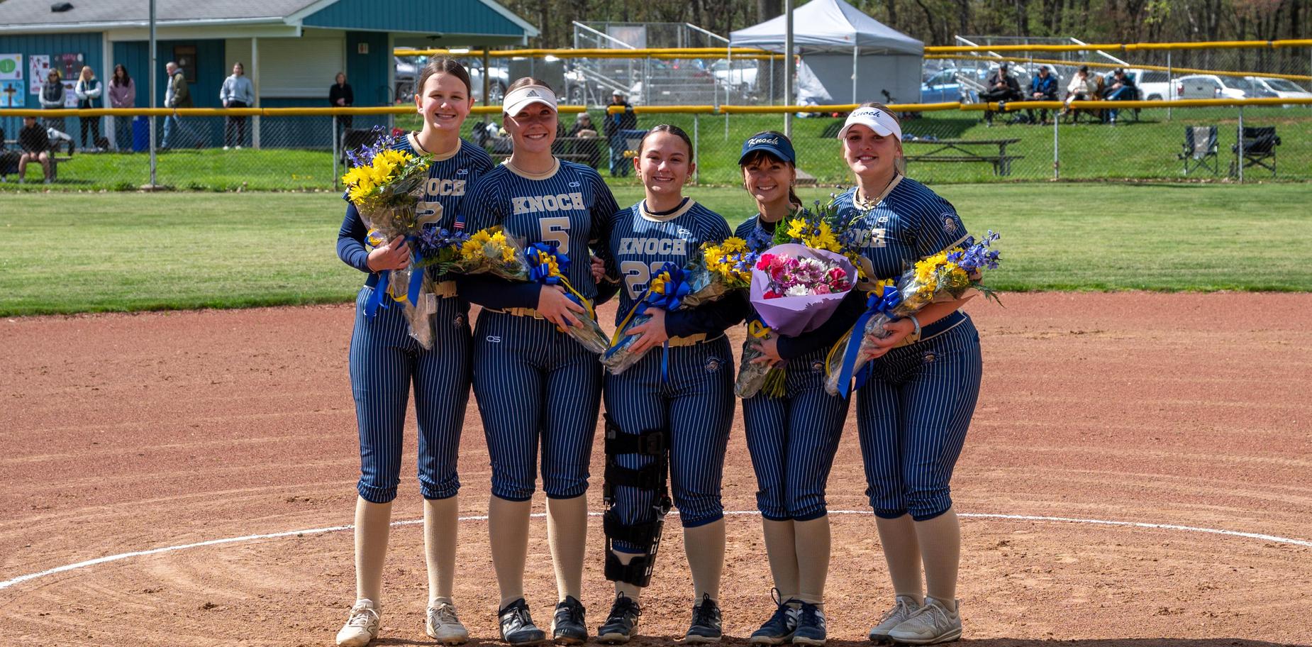 Five female softball players posing with bouquets and awards on a field.