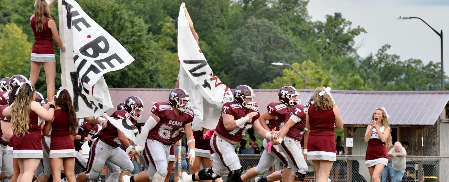 Football team runs onto the field, holding large banners with cheerleaders in the background.