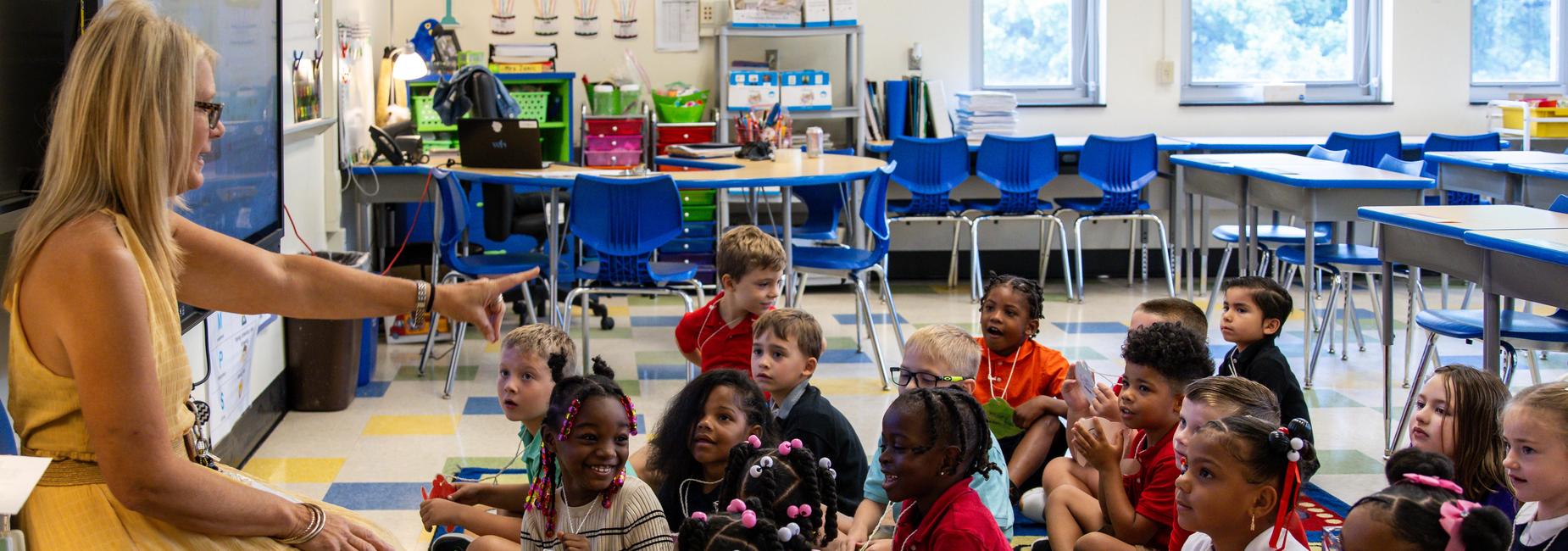 An elementary teacher sits in front of seated students, who are all smiling and laughing