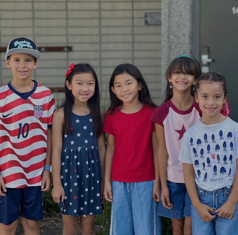 Five children posing together outdoors, wearing casual summer outfits.