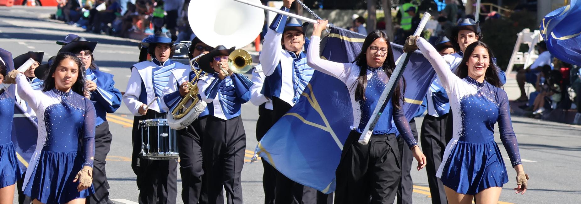 A marching band in blue uniforms parades with flags and musical instruments.