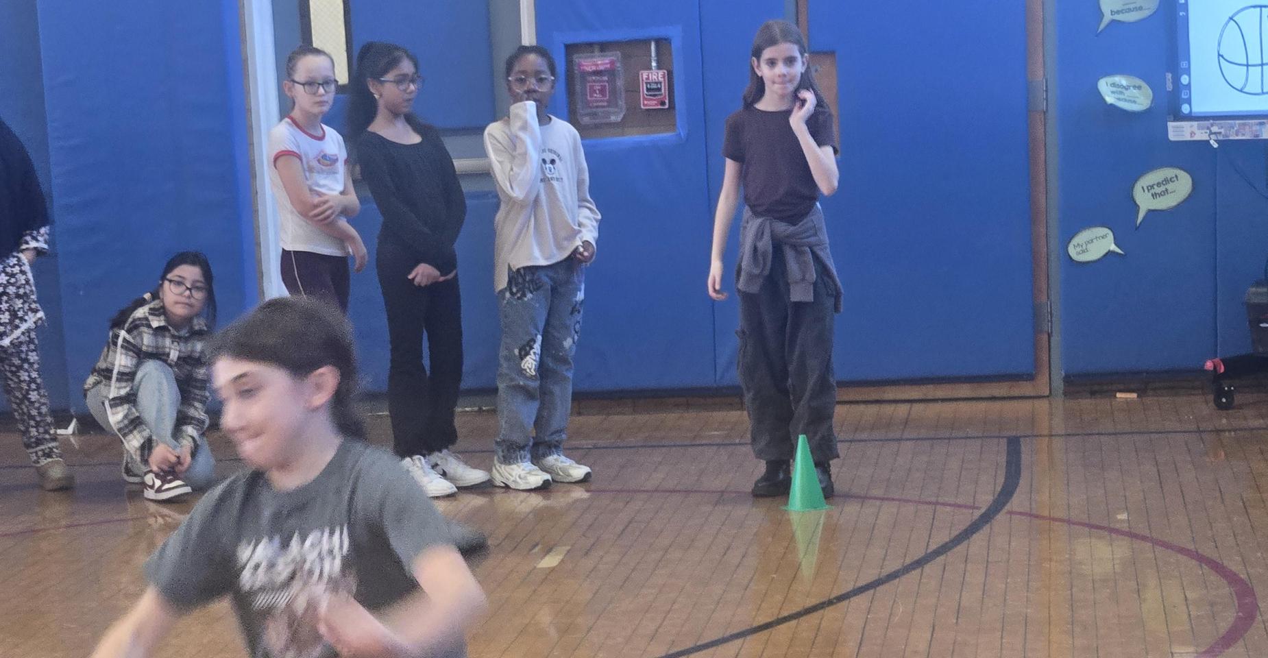 A group of children in a gym, some waiting and one running towards a cone.