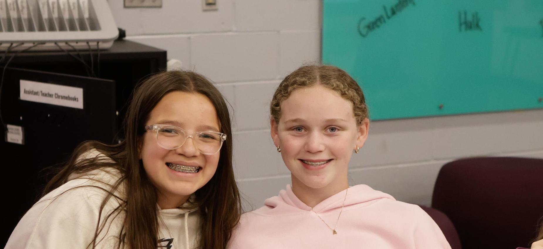 Two young girls smiling together in a classroom setting.