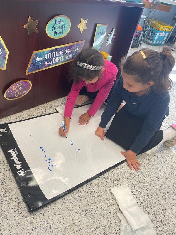 Two third grade girls spread their white board on the ground to complete a math problem together.