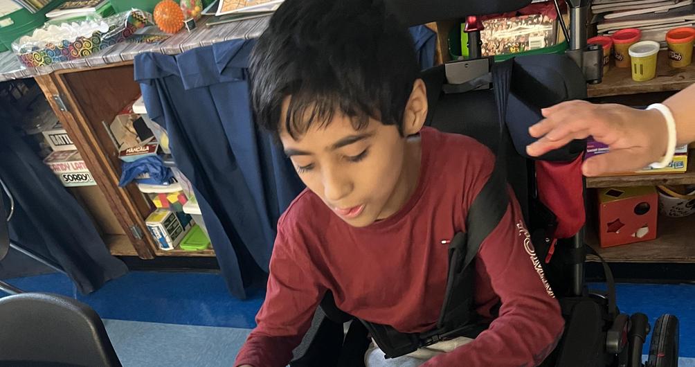 Child in a wheelchair decorating a mask with paint and glue on a table.