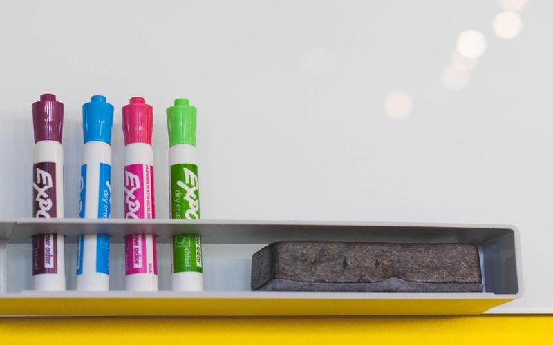 Four colorful dry erase markers and an eraser on a whiteboard shelf.