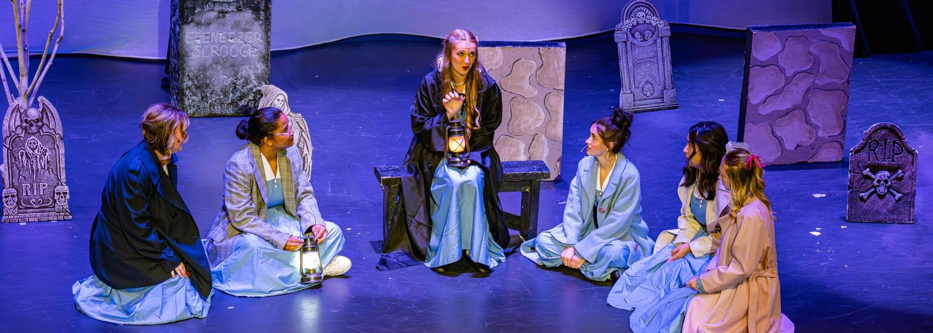 A group of six performers in costumes seated on stage around a storyteller with props.