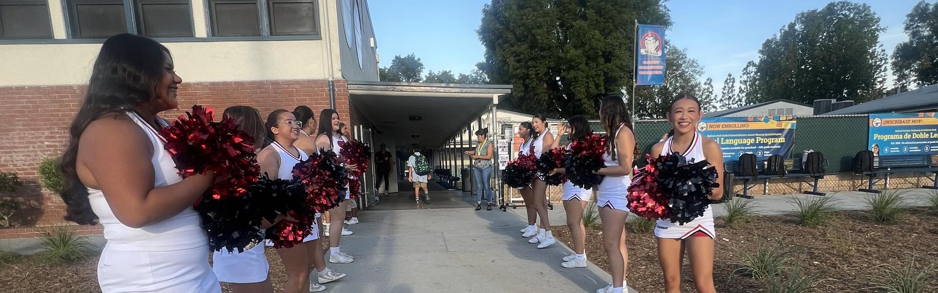 Cheerleaders in white uniforms holding pom-poms, forming a line on a school pathway.