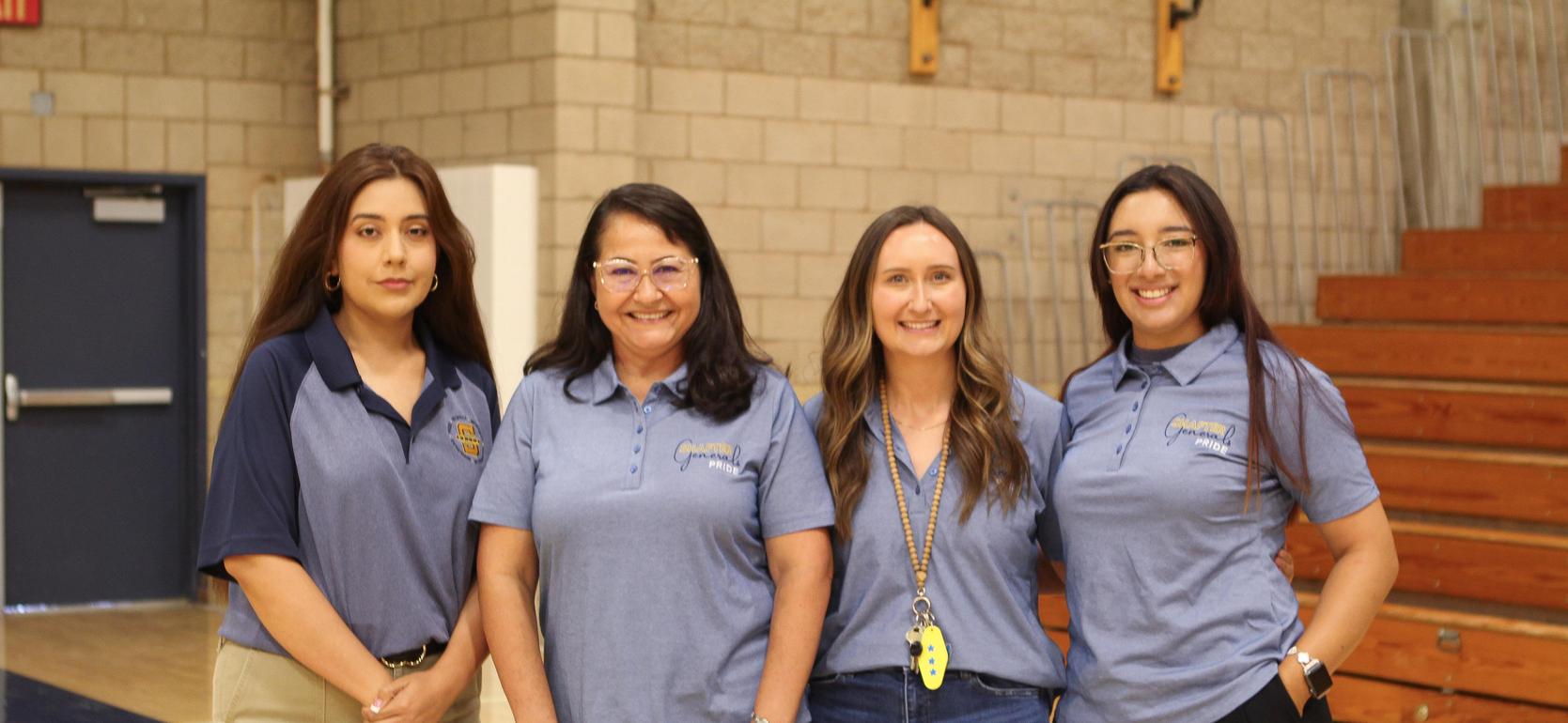 Four women in blue and black clothing pose together in a gym with wooden bleachers.