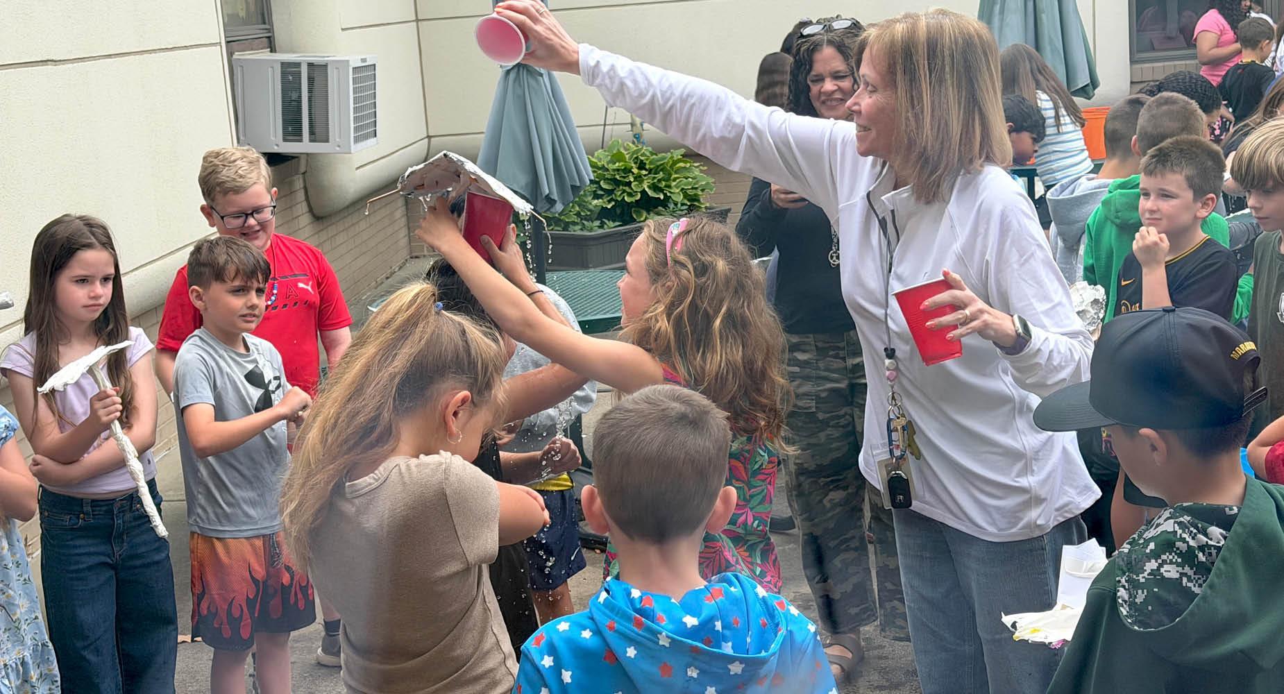 A teacher dumps a cup of water over a student's head while she holds a handmade umbrella
