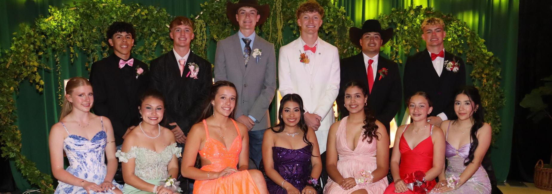 Formal gathering of students in elegant dresses and suits, posing in front of greenery.