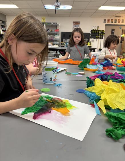 A young girl paints on paper with colorful materials around her at a creative workspace.