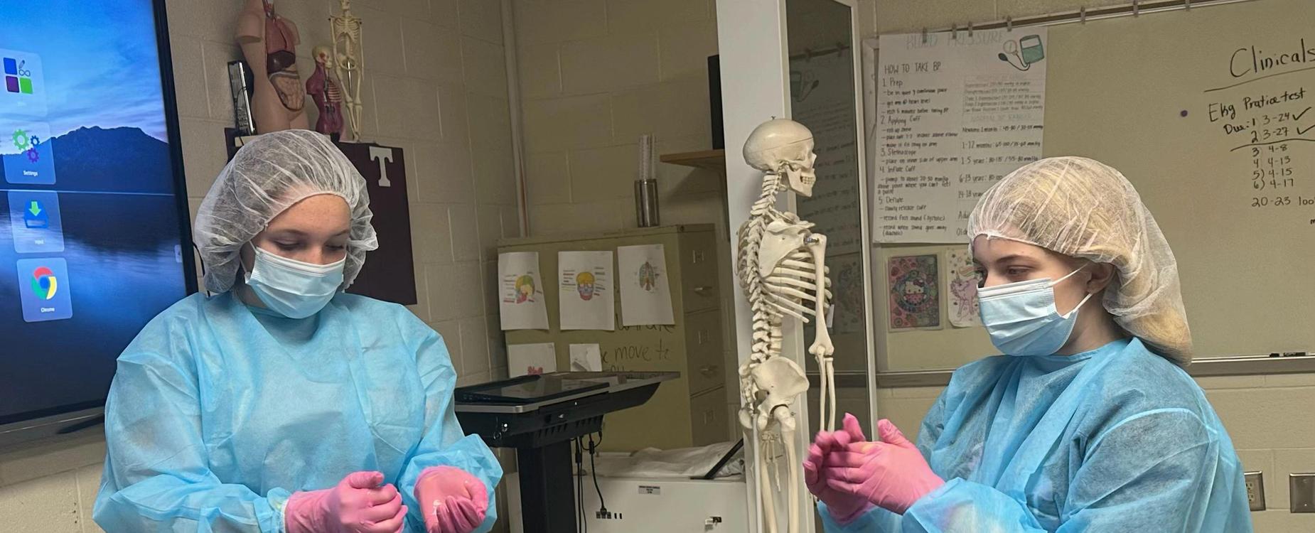 Two students in protective gear preparing for an activity near a skeleton model.
