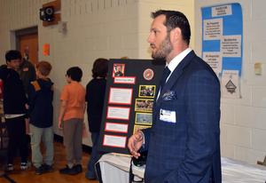 A man in a suit speaks near a presentation board with students in the background.