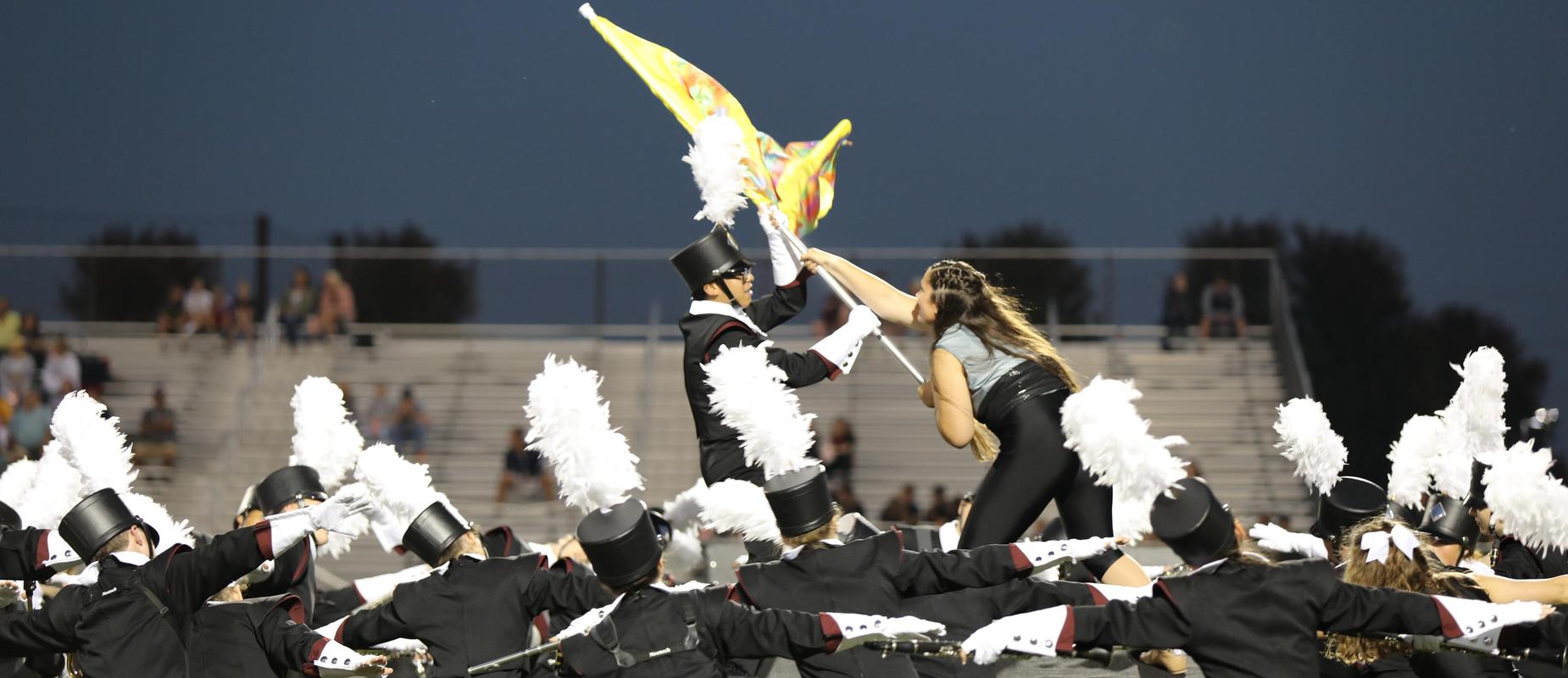 marching band students encircle two who are grabbing band flag on field